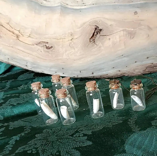 Five small clear glass vials with cork stoppers, each containing a white feather, arranged diagonally on a dark green patterned fabric next to a large natural wood slab.